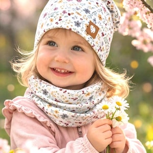 Könnte beinhalten: Ein Kind trägt eine weiße Mütze und einen Schal mit Blumenmuster. Das Kind hält einen kleinen Strauß Gänseblümchen und trägt eine rosa Jacke. Auf der Mütze befindet sich ein brauner Lederaufnäher.