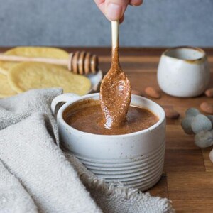 May include: A white ceramic mug filled with a thick, brown almond butter. A spoon with almond butter is being dipped into the mug.