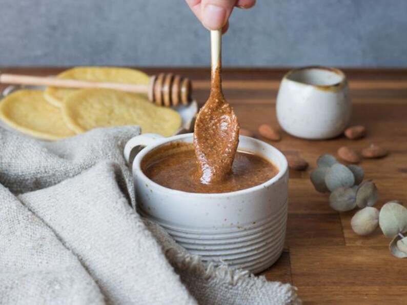 May include: A white ceramic mug filled with a thick, brown almond butter. A spoon with almond butter is being dipped into the mug.