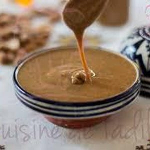 May include: A close-up of a bowl of tahini, a creamy sesame seed paste, with a dollop of tahini being drizzled from a spoon. The bowl is decorated with a blue and white pattern.