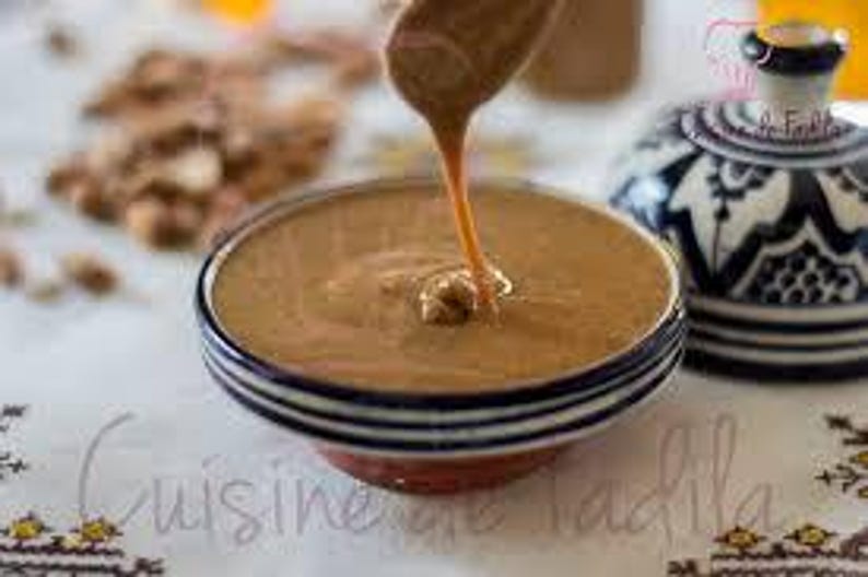 May include: A close-up of a bowl of tahini, a creamy sesame seed paste, with a dollop of tahini being drizzled from a spoon. The bowl is decorated with a blue and white pattern.