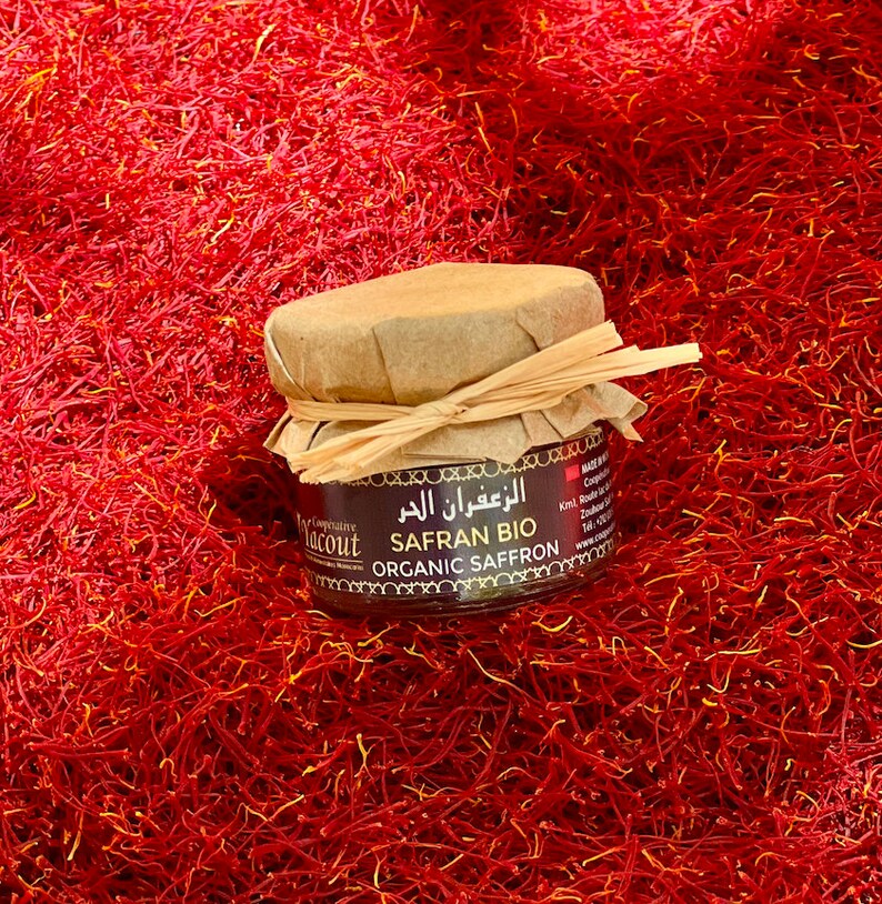 May include: A small glass jar filled with organic saffron threads. The jar has a brown paper label with the text "Safran Bio Organic Saffron" in English and Arabic. The jar is sitting on a bed of red saffron threads.