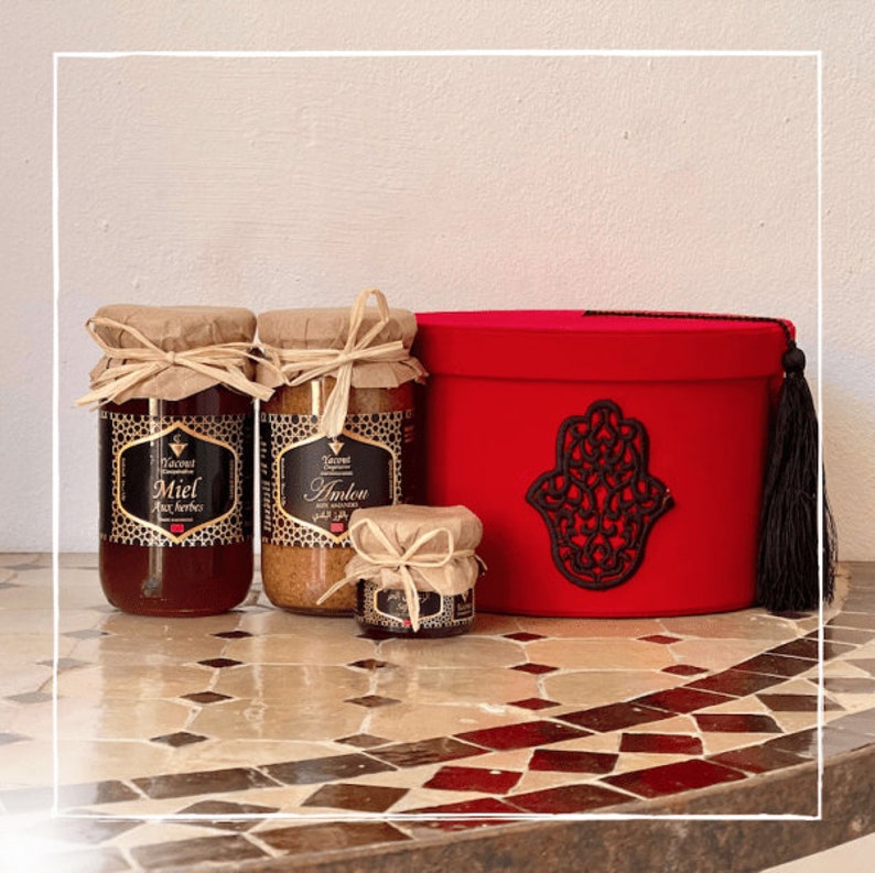 May include: A red velvet gift box with a black Hamsa hand design and a black tassel. The box contains three glass jars of honey, one labeled "Miel aux Herbes" and another labeled "Amlou".