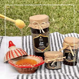 May include: Three glass jars of honey with brown paper lids and black labels. The labels have Arabic and French text that reads "Miel aux Herbes de Souk" and "Honey with Sour Herbs". The jars are on a black and white striped surface with a wooden honey dipper and a red and white ceramic bowl.