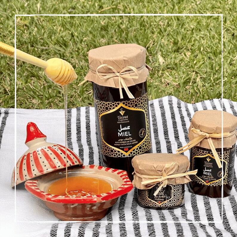 May include: Three glass jars of honey with brown paper lids and black labels. The labels have Arabic and French text that reads "Miel aux Herbes de Souk" and "Honey with Sour Herbs". The jars are on a black and white striped surface with a wooden honey dipper and a red and white ceramic bowl.