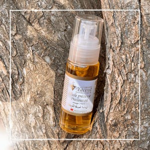 May include: A clear glass bottle of precious oil with a pump dispenser. The bottle has a white label with text in multiple languages, including "Precious oil" and "Visage / face." The oil is amber-colored. The bottle is leaning against a tree trunk.