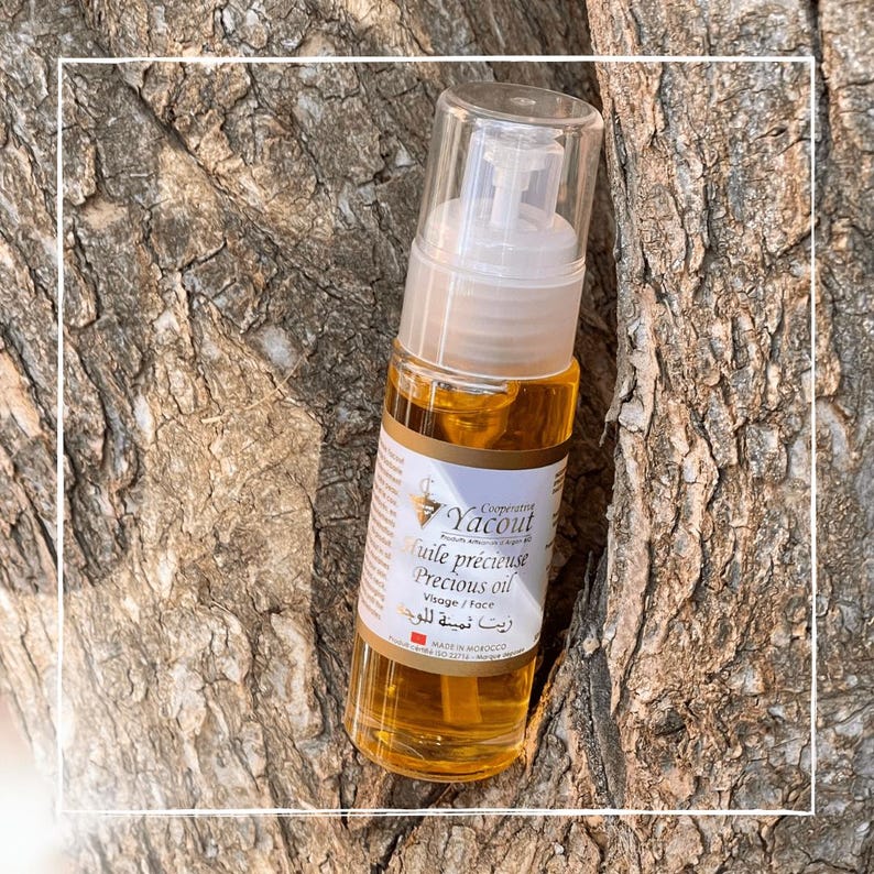May include: A clear glass bottle of precious oil with a pump dispenser. The bottle has a white label with text in multiple languages, including "Precious oil" and "Visage / face." The oil is amber-colored. The bottle is leaning against a tree trunk.