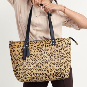 May include: A tan and black leopard print tote bag with black handles and a tassel. The bag is held by a person. The bag has a zipper closure and is made of a textured material.