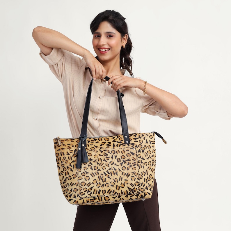 May include: A tan and black leopard print tote bag with black handles and a tassel. The bag is held by a person. The bag has a zipper closure and is made of a textured material.