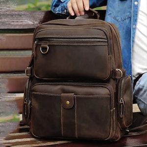 Pode incluir: Uma mochila de couro marrom com vários bolsos e fecho de correr. A mochila tem um aspeto envelhecido e apresenta uma fivela de latão.