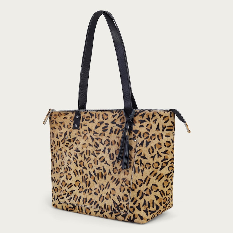 Peut inclure: Un sac fourre-tout rectangulaire avec un motif l&eacute;opard en tons de beige et noir. Il est dot&eacute; de poign&eacute;es en cuir noir et d'un pompon noir. Une fermeture &eacute;clair parcourt le bord sup&eacute;rieur.
