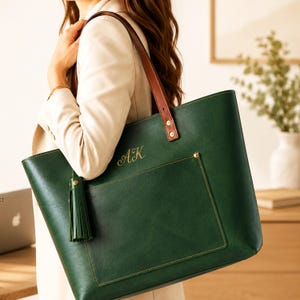Peut inclure: Un sac fourre-tout vert foncé avec des anses en cuir marron, une grande poche avant et un pompon assorti. Le sac est personnalisé avec les initiales AK brodées en doré sur le devant. Le design présente des détails de coutures visibles.
