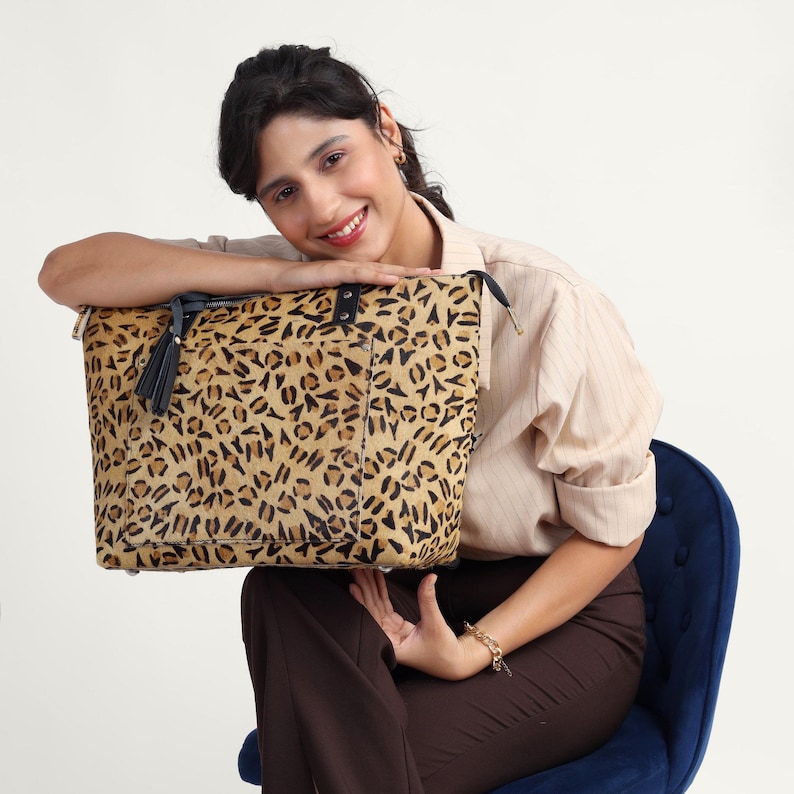 May include: A leopard print tote bag with a black top and a black tassel. The bag is held by a person wearing a beige striped shirt and brown pants. The bag has a black handle and a gold zipper. The person is sitting on a blue chair.
