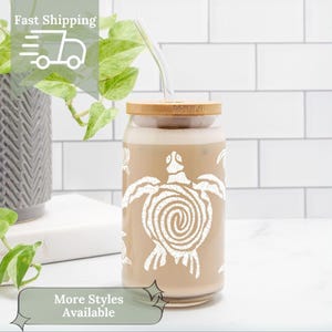 May include: A clear glass tumbler with a bamboo lid and a white sea turtle design. The tumbler contains a light brown liquid and a clear glass straw. The words "More Styles Available" are at the bottom of the image.