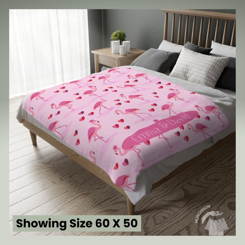 Adult Minky Blanket Flamingo Decor Couch Throw with a blanket with flamingos and hearts printed all over and name personalization area laying across the foot of a bed. Tag at bottom reads showing size 60x50.