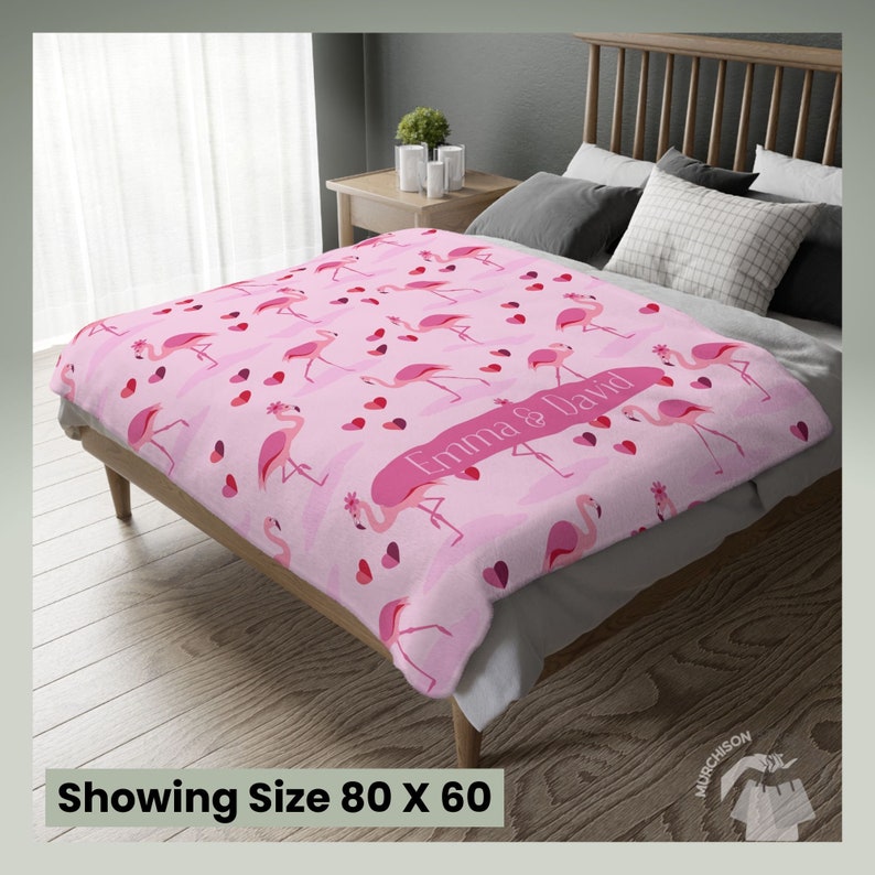 Couch Throw Blanket Valentine Gift with a blanket with flamingos and hearts printed all over and name personalization area laying across the foot of a bed. Tag at bottom reads showing size 80x60.