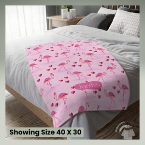Love Throw Blanket Adult Minky Blanket Flamingo Decor with a blanket with flamingos and hearts printed all over and name personalization area laying across the foot of a bed. Tag at bottom reads showing size 40x30.