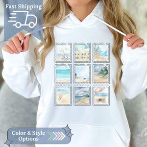 Può includere: Felpa con cappuccio bianca con una griglia di nove disegni di francobolli a tema spiaggia. Ogni francobollo raffigura una scena acquerellata diversa di una spiaggia con ombrelloni, palme e vista sull'oceano. La felpa ha un cappuccio con coulisse.