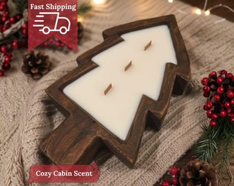 May include: A wooden Christmas tree-shaped candle holder filled with white wax and three wooden wicks. The candle sits on a knitted surface, surrounded by pine cones and red berries. The text "Cozy Cabin Scent" is visible.