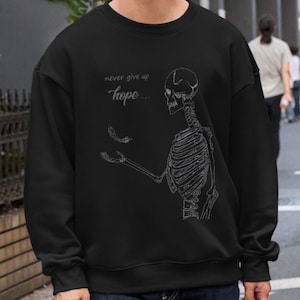 以下が含まれることがあります： 前面に白い骸骨のグラフィックと「never give up hope...」というテキストがプリントされた黒いスウェットシャツです。