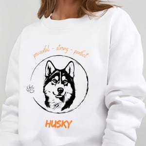 Könnte beinhalten: Weißes Sweatshirt mit einer schwarz-weißen Illustration eines Husky-Hundes in einem Kreis. Die Worte "powerful - strong - perfect" stehen über dem Hund, und das Wort "HUSKY" steht darunter in Orange.