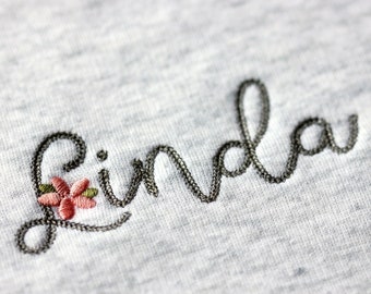 May include: Embroidered name "Linda" in black thread on a gray fabric background with a small pink flower.