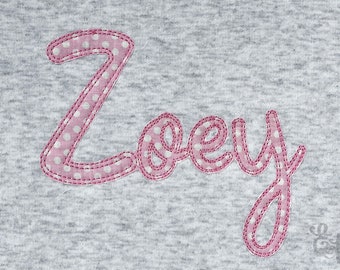 May include: Pink fabric with white polka dots sewn into the name "Zoey" in a cursive font.