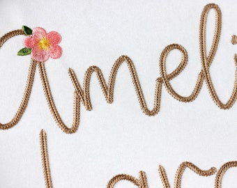 May include: Embroidered name "Amelia" in a cursive font with a pink flower and green leaves above the first letter.