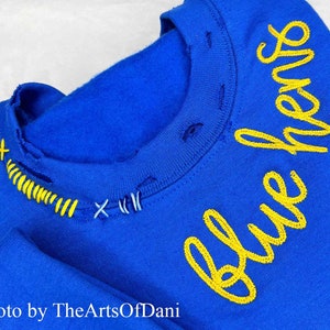 May include: Royal blue sweatshirt with yellow embroidered text that reads "blue hens". The neckline has been distressed and stitched with yellow and blue thread. The photo is credited to TheArtsOfDani.