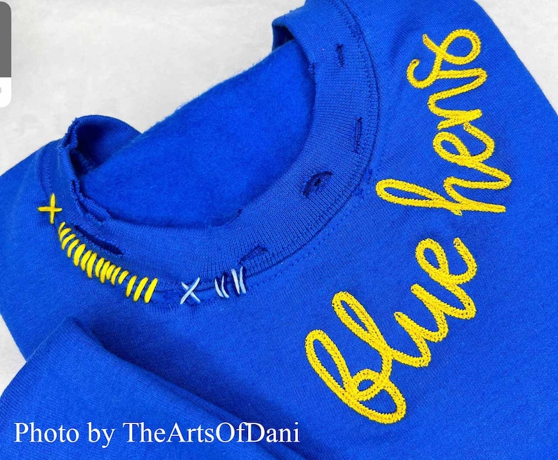 May include: Royal blue sweatshirt with yellow embroidered text that reads "blue hens". The neckline has been distressed and stitched with yellow and blue thread. The photo is credited to TheArtsOfDani.
