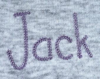 May include: A close-up of the word "Jack" embroidered in purple thread on a gray fabric.