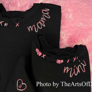 May include: Black sweatshirts with pink and white embroidered details. One sweatshirt says "mama" and the other says "mini". The neckline has pink cross-stitch accents. A pink heart with a "B" is embroidered on one sweatshirt. Photo by TheArtsOfDani.