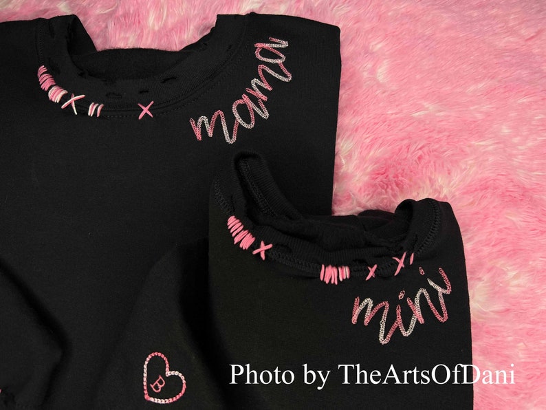 May include: Black sweatshirts with pink and white embroidered details. One sweatshirt says "mama" and the other says "mini". The neckline has pink cross-stitch accents. A pink heart with a "B" is embroidered on one sweatshirt. Photo by TheArtsOfDani.