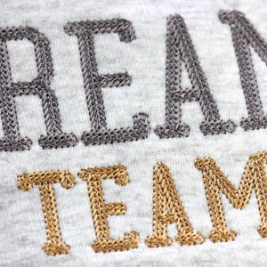 May include: Gray embroidered "DREAM" text above gold embroidered "TEAM" text on a gray fabric.