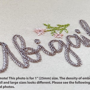 May include: Close-up of the name "Abigail" embroidered in a silver-gray thread on a light beige fabric. Delicate pink and green floral embroidery accents the name. The text below states the photo is for a 1-inch size and details about the embroidery.