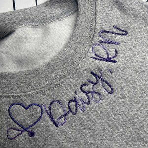 May include: A gray crewneck sweatshirt with embroidered purple text and a heart-shaped stethoscope design. The text reads "Daisy, RN". The sweatshirt is made of a soft material and is suitable for casual wear.