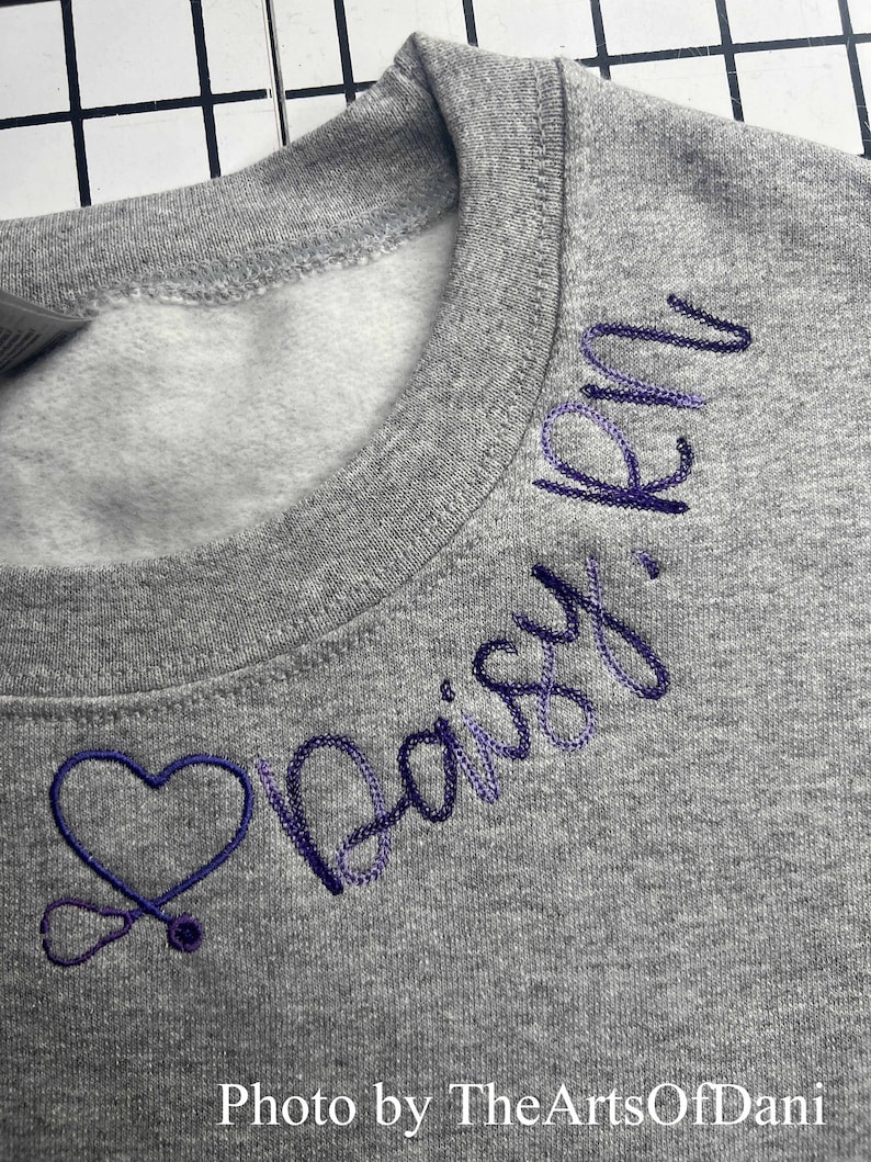 May include: A gray crewneck sweatshirt with embroidered purple text and a heart-shaped stethoscope design. The text reads "Daisy, RN". The sweatshirt is made of a soft material and is suitable for casual wear.