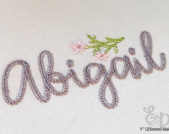 May include: Embroidered name "Abigail" in silver thread with floral accents. The script is stitched on a beige fabric, with a sprig of green leaves and pink flowers. The text is approximately 1 inch (25mm) in size.