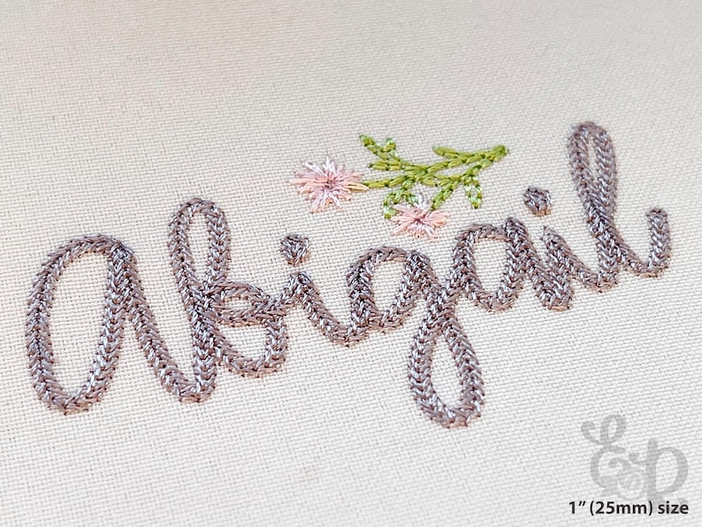 May include: Embroidered name "Abigail" in silver thread with floral accents. The script is stitched on a beige fabric, with a sprig of green leaves and pink flowers. The text is approximately 1 inch (25mm) in size.
