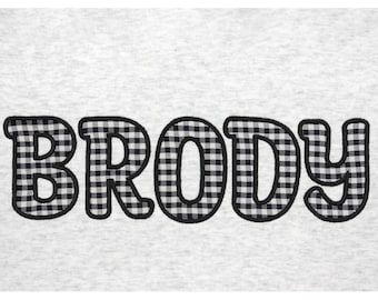 Puede incluir: Una sudadera gris con el nombre "Brody" bordado en vichy blanco y negro.
