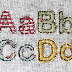 Peut inclure: Gros plan sur les lettres en tissu A, a, B, b, C, c, D et d, chacune avec un motif unique et une bordure cousue. Les lettres sont de couleurs variées, dont des rayures rouges et blanches, des motifs floraux et du vichy jaune.