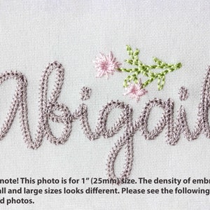 May include: Embroidered name "Abigail" in a cursive font, stitched in a light mauve thread. Delicate pink flowers and green leaves are embroidered above the "ail" letters. The text is on a white fabric background.