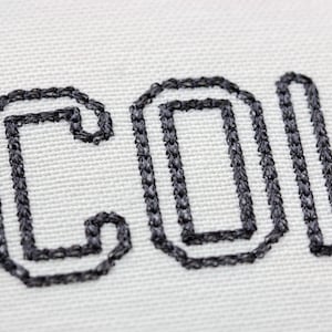 May include: Close-up of white fabric with the embroidered word "COL" in dark gray thread. The letters are outlined with a thick, textured stitch, creating a bold and tactile appearance. The fabric has a subtle, woven texture.