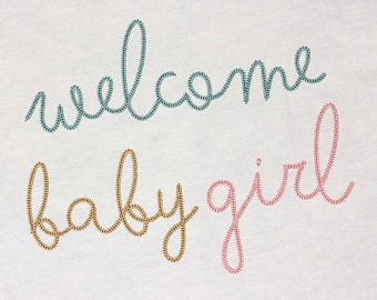 May include: Welcome baby girl embroidered on a white fabric background. The words are in a cursive font. The word "welcome" is in teal blue thread, the word "baby" is in gold thread, and the word "girl" is in pink thread.