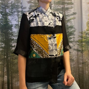 Op de afbeelding: Een patchwork shirt met een uniek design. Het shirt heeft een mix van zwart, wit, geel en groen bedrukte stoffen. Het heeft korte mouwen met zwarte satijnen bies en een mandarijnkraag. Het shirt is dichtgeknoopt en lijkt uit meerdere materialen te bestaan.