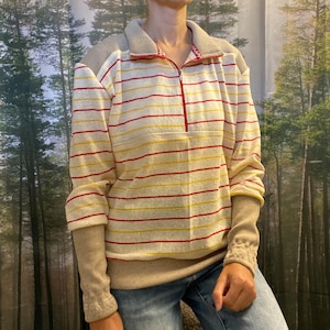 Könnte beinhalten: Ein Langarm-Pullover mit halbem Reißverschluss, einer cremefarbenen Basis mit horizontalen roten und gelben Streifen sowie beigefarbenem Rippstrick an Schultern, Ärmelbündchen und Saum. Die Krageninnenseite zeigt ein kleines rot-weißes Muster.