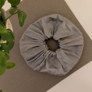 May include: A grey scrunchie made from fabric with a gathered design. The scrunchie is circular with a hole in the centre. The fabric has a subtle sheen. The scrunchie is on a grey surface.