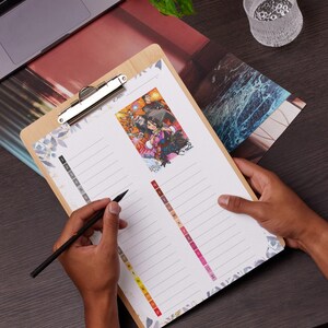 May include: A wooden clipboard with a silver clip holding a lined paper with a colourful illustration. The paper has a floral design on the edges and a series of coloured tabs. A black pen is held over the paper.