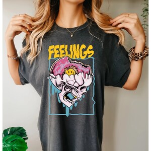May include: Dark gray t-shirt with the word "FEELINGS" in yellow above a graphic of a skull with a pink donut brain and a smiling face. The skull is outlined in blue.