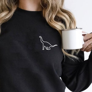 Peut inclure: Sweat-shirt noir avec un contour blanc de dinosaure sur la poitrine gauche. Le sweat-shirt à col rond est noir. Une tasse blanche avec un bord argenté est tenue dans la main droite. Style décontracté.